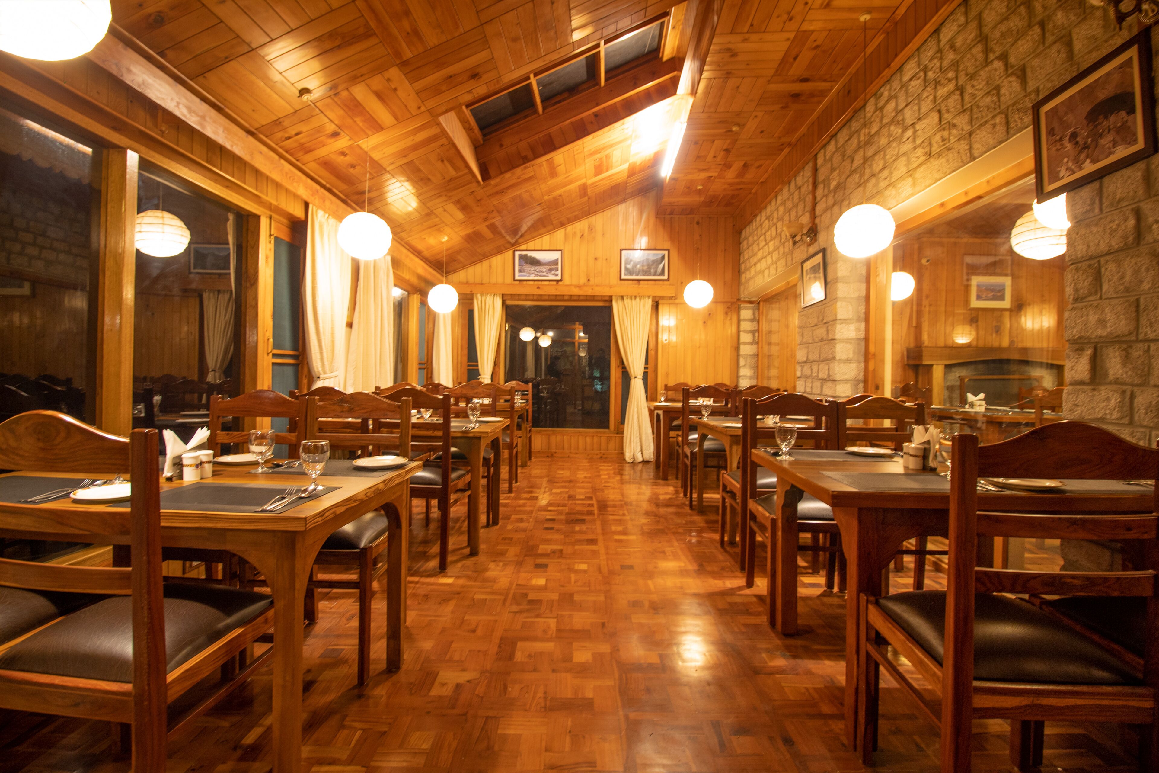 A restaurant with wooden tables and chairs, a stone wall, and hanging lights.