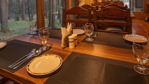 A dining table set with plates, cutlery, and glasses, with a view of a forest through large windows. - Hotel  Negis Mayflower  (Manali)