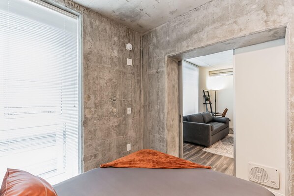 A room with a concrete wall, a bed with an orange pillow, a grey sofa, and a lamp in the background.