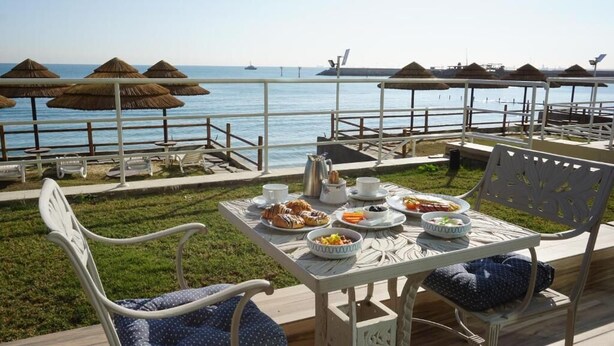 Uma mesa de jantar com pratos de comida, uma cafeteira e uma tigela de frutas, em um deck com vista para o mar e guarda-sóis de palha.