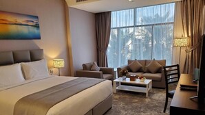 Quarto de hotel com uma cama grande, um sofá, uma mesa de centro e uma escrivaninha. Há cortinas, um quadro na parede e uma janela com vista.