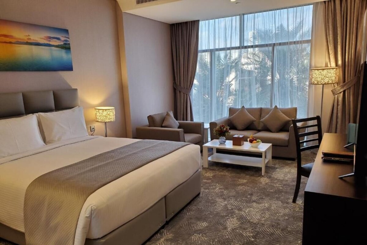 Een hotelkamer met een groot bed, een bank, een salontafel en een bureau. Gordijnen, een schilderij aan de muur en een raam met uitzicht.