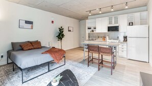 Un apartamento moderno con cama, una pequeña mesa de comedor, cocina con armarios blancos y una planta.