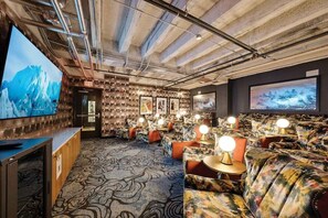 Ein gemütlicher Lounge-Bereich mit Sofas, Tischen, einem großen Bildschirm und einem gemusterten Teppich.