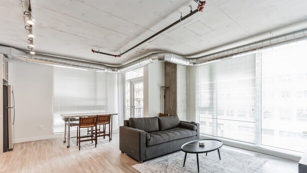 Una sala de estar moderna con un sofá gris, una mesa redonda de centro y una mesa de comedor con sillas. Hay ventanas grandes con persianas, un techo con conductos a la vista y una lámpara.