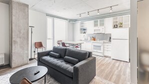 Una sala moderna con un sofá gris, una mesa de centro y un comedor con mesa y sillas. Se ve la cocina, con gabinetes y electrodomésticos blancos.