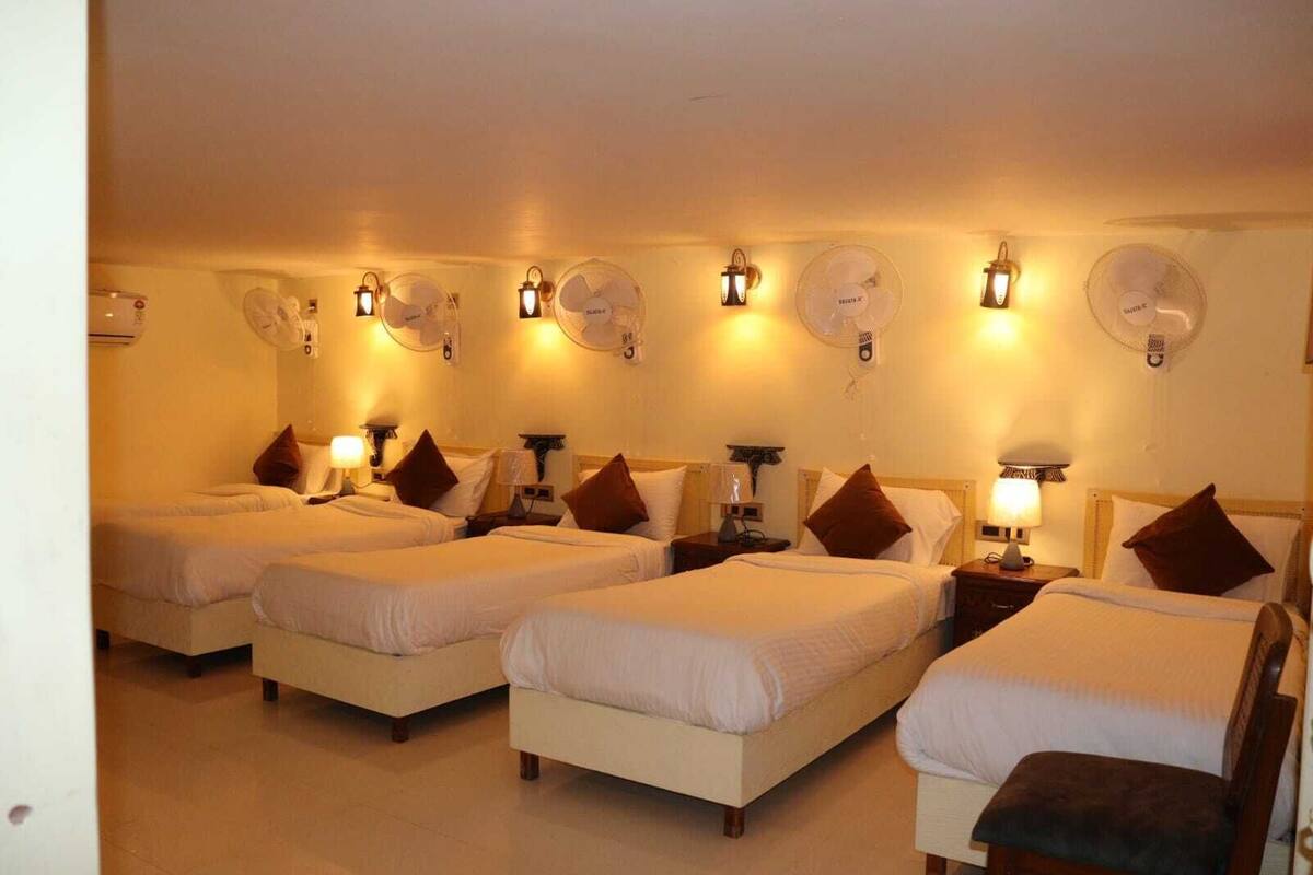 Una habitació d'hotel amb quatre llits individuals, cadascun amb una taula de nit i una làmpada. Hi ha ventiladors fixats a la paret sobre cada llit.