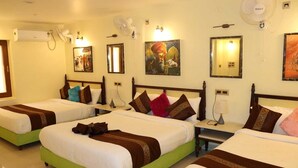 A hotel room with three single beds, each with a green base and white bedding. There are wall-mounted fans and a painting above the beds. - Satshree Kothi (Varanasi)