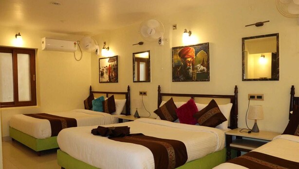 A hotel room with two single beds, a green accent bed, a nightstand with a lamp, and a wall-mounted fan. - Satshree Kothi (Varanasi)
