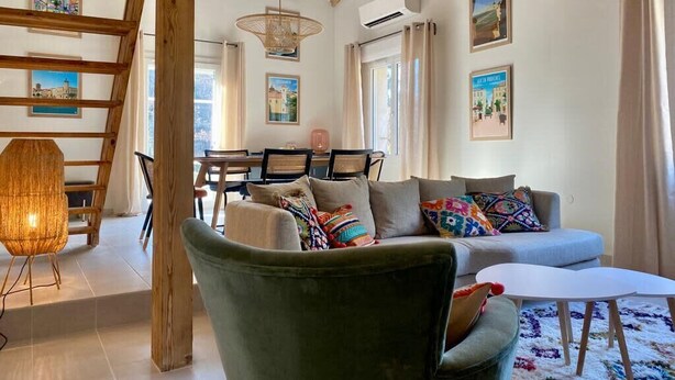 Un salon chaleureux avec un fauteuil vert, un canapé et une petite table blanche. On y trouve une structure de poutres apparentes en bois, un lampadaire en osier et un coin repas avec des chaises et une table.