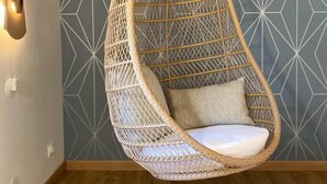 Une chaise suspendue en rotin avec un coussin, des poutres en bois et un mur orné d’un motif géométrique.