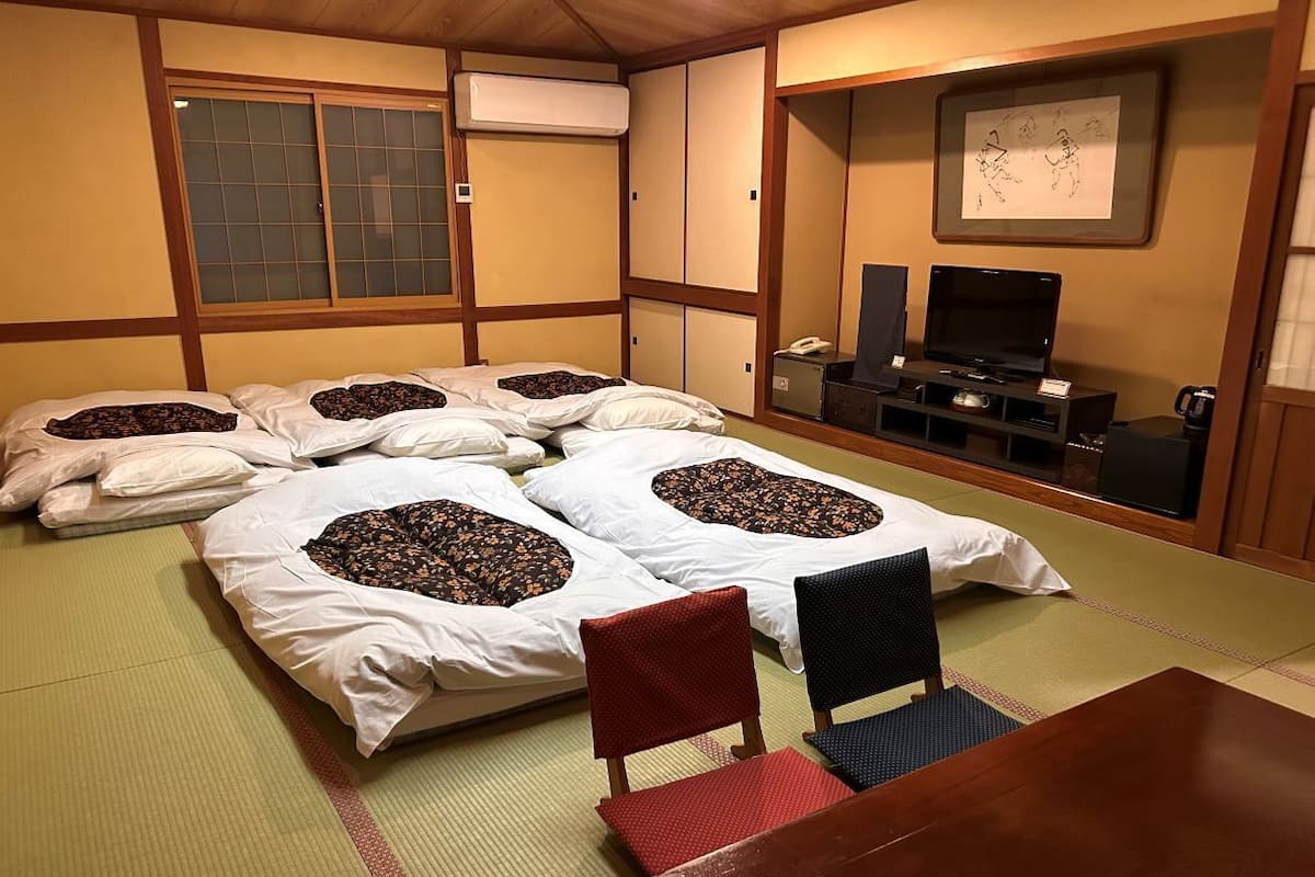 A traditional Japanese room with tatami mats, a tatami mat table, two tatami mat beds with pillows, a television, and a small cabinet.