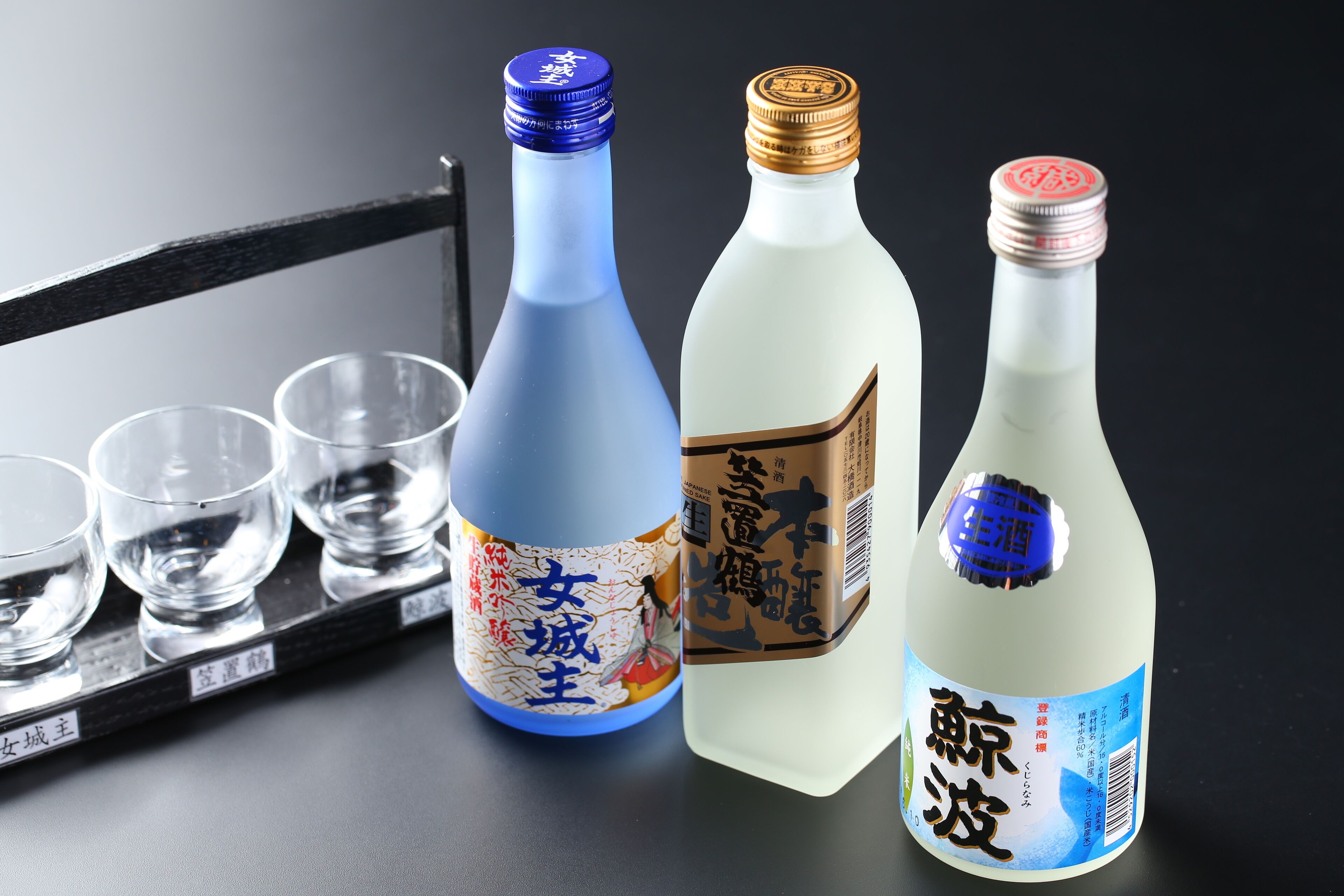 Three bottles of sake with labels and four empty glasses on a black surface.