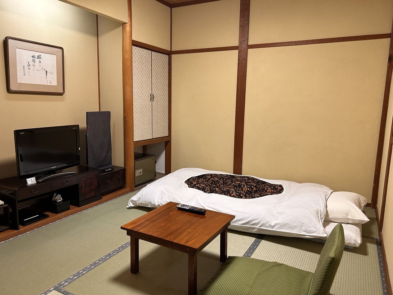 Una habitación tradicional japonesa con suelo de tatami, una cama con almohada, un televisor, un armario y una mesita.