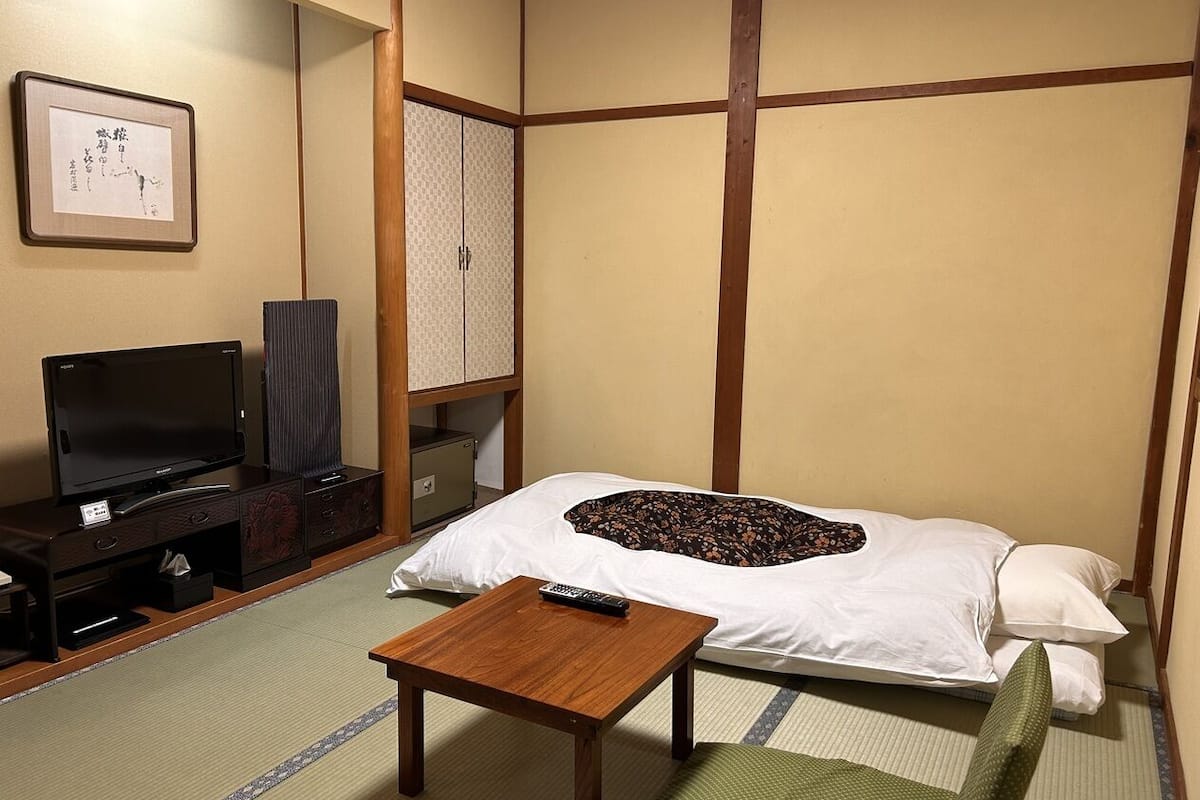 A traditional Japanese room with tatami flooring, a bed with a pillow, a television, a cabinet, and a small table.