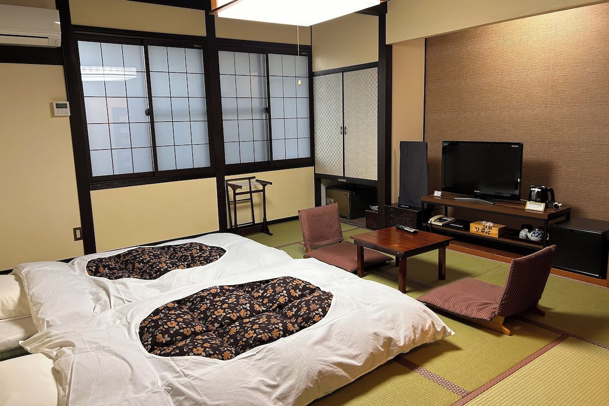 A traditional Japanese-style hotel room with tatami flooring, a bed with floral-patterned bedding, a television, and a small table with a remote control.