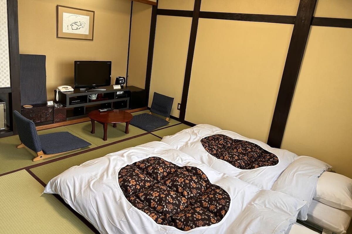 A traditional Japanese-style room with tatami flooring, a low table, a sofa, and a bed with floral-patterned pillows.