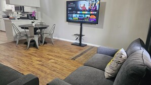 A modern living room with a gray sofa, a dining table with chairs, a flat-screen TV mounted on a stand, and a kitchen area in the background. - ENTIRE UNIT | 2BR | Washer/Dryer | Sleeps 6 | Near Airport (Austin)