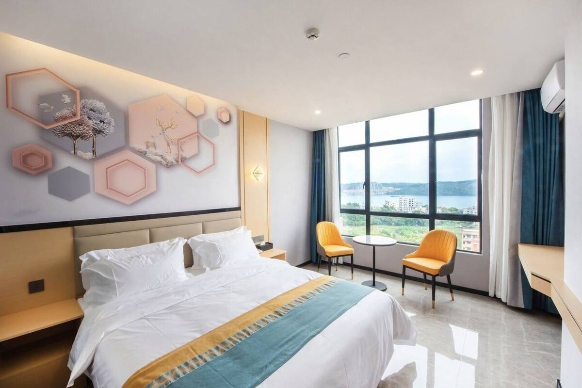 Una habitación de hotel con una cama grande, dos sillas, una mesita y vistas a la ciudad a través de amplias ventanas.