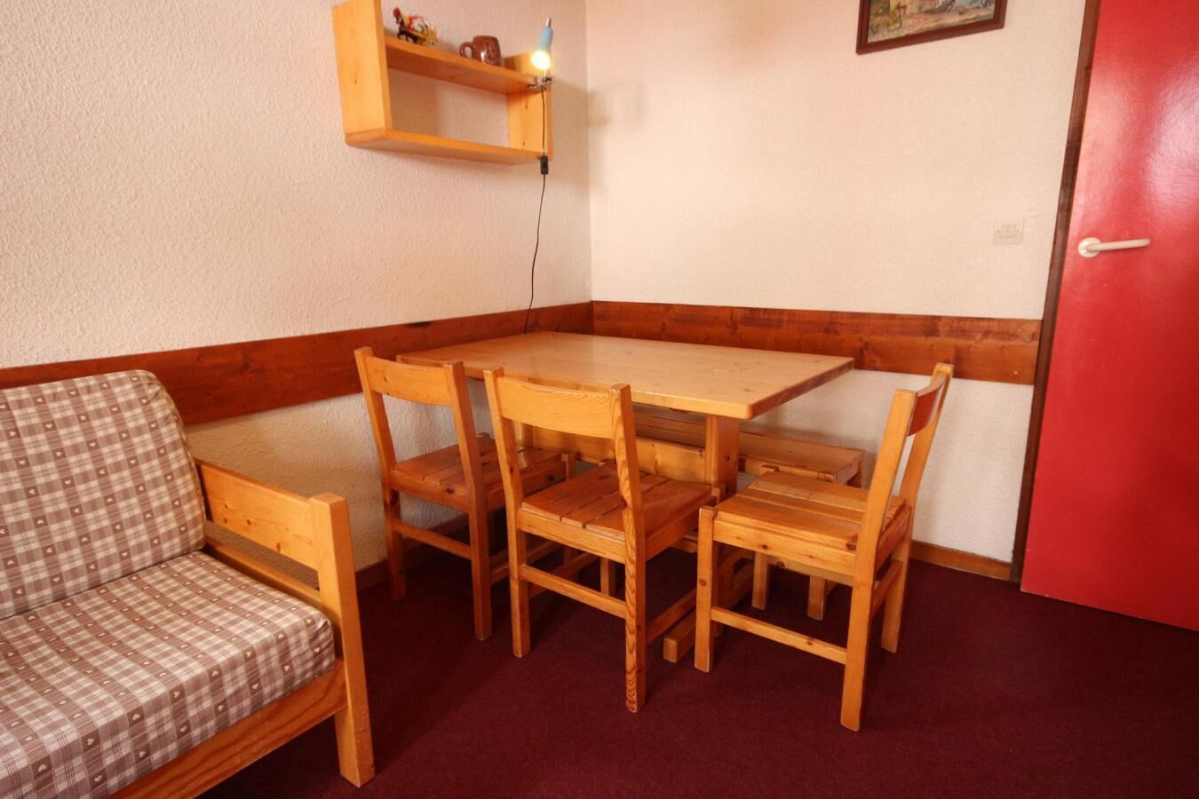 A corner room with a wooden table and four chairs, a plaid sofa, a red door, and a wall-mounted shelf with a lamp.