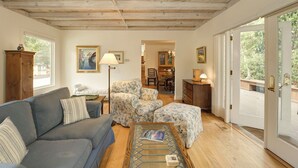 Una sala de estar con sofá, sillón, reposapiés y mesa de centro. Tiene techo de madera, una puerta que da al exterior y un área de comedor al fondo.