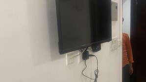 A flat-screen TV mounted on a white wall with a power cord hanging below it. - Hotel Avalon Palace  (New Delhi)
