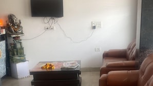 A living room with a brown leather sofa, a coffee table with magazines, a television mounted on the wall, and a ceiling fan. - Hotel Avalon Palace  (New Delhi)