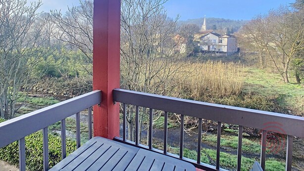 Un balcon avec un poteau rouge et une balustrade en métal, donnant sur un paysage rural avec des maisons et des arbres.
