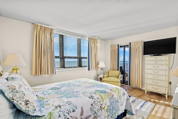 A bedroom with a bed, curtains, a TV, a dresser, and a view of the sea. - Oceanfront Condo In Quiet, Secured Building + Free Attraction Tickets! (Myrtle Beach)