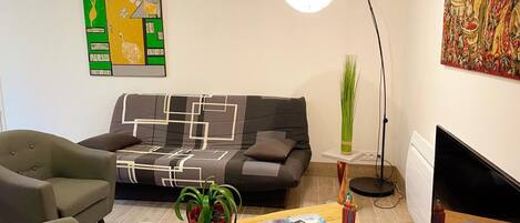 Una sala de estar moderna con un sofá gris, un sillón verde, una mesa de centro de madera con revistas, una planta en maceta y una lámpara de pie.