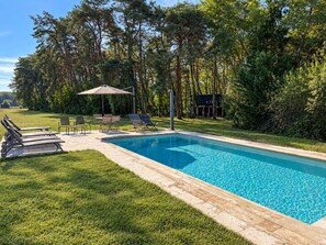A rectangular swimming pool with a tiled edge, surrounded by a grassy area with lounge chairs and a table under a canopy.