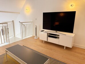 A modern living room with a flat-screen TV on a white cabinet, a standing heater, and a staircase with white railings. - Luxury Lodge in Fontaines-en-Sologne: Heated Pool, Private Lake, Activities & Scenic Views (Fontaines-en-Sologne)