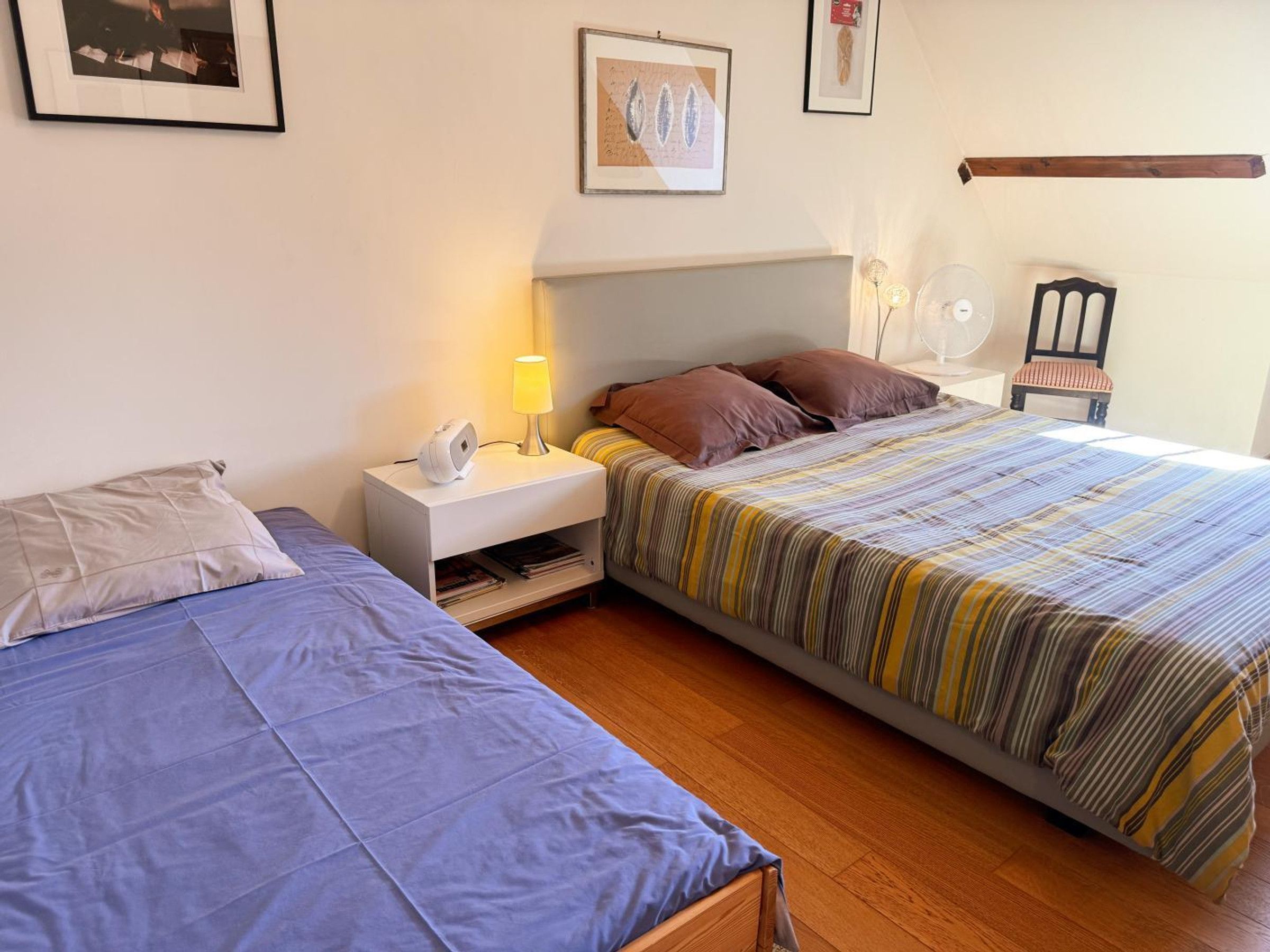 Una habitación de hotel con dos camas, una mesita de noche con lámpara, una silla y un ventilador de pared.