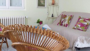 Una habitación con una silla de mimbre, un sofá con cojines estampados, un radiador y una estantería con un jarrón.