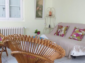 A room with a wicker chair, a sofa with patterned cushions, a radiator, and a wall-mounted shelf with a vase. - Family cottage near Loire castles, pets welcome, private garden (Saint-Gervais-la-Forêt)