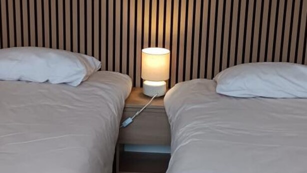 Habitación de hotel con dos camas, una mesita de noche con lámpara y cabecero de madera con listones.