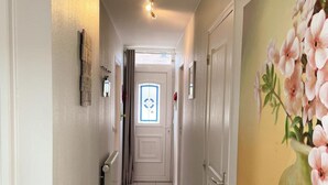Un pasillo con una puerta al final, una ventana con diseño de rombos y un cuadro de flores en la pared.