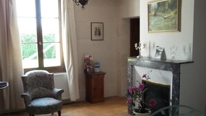 A room with a fireplace, a chair, a window with curtains, and a painting on the wall. - Restored Country House with WIFI and Fireplaces (Prunay-Cassereau)