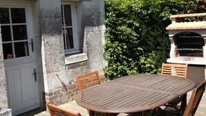A stone building with a white door and windows, a wooden table and chairs, and a brick oven in the background. - Restored Country House with WIFI and Fireplaces (Prunay-Cassereau)