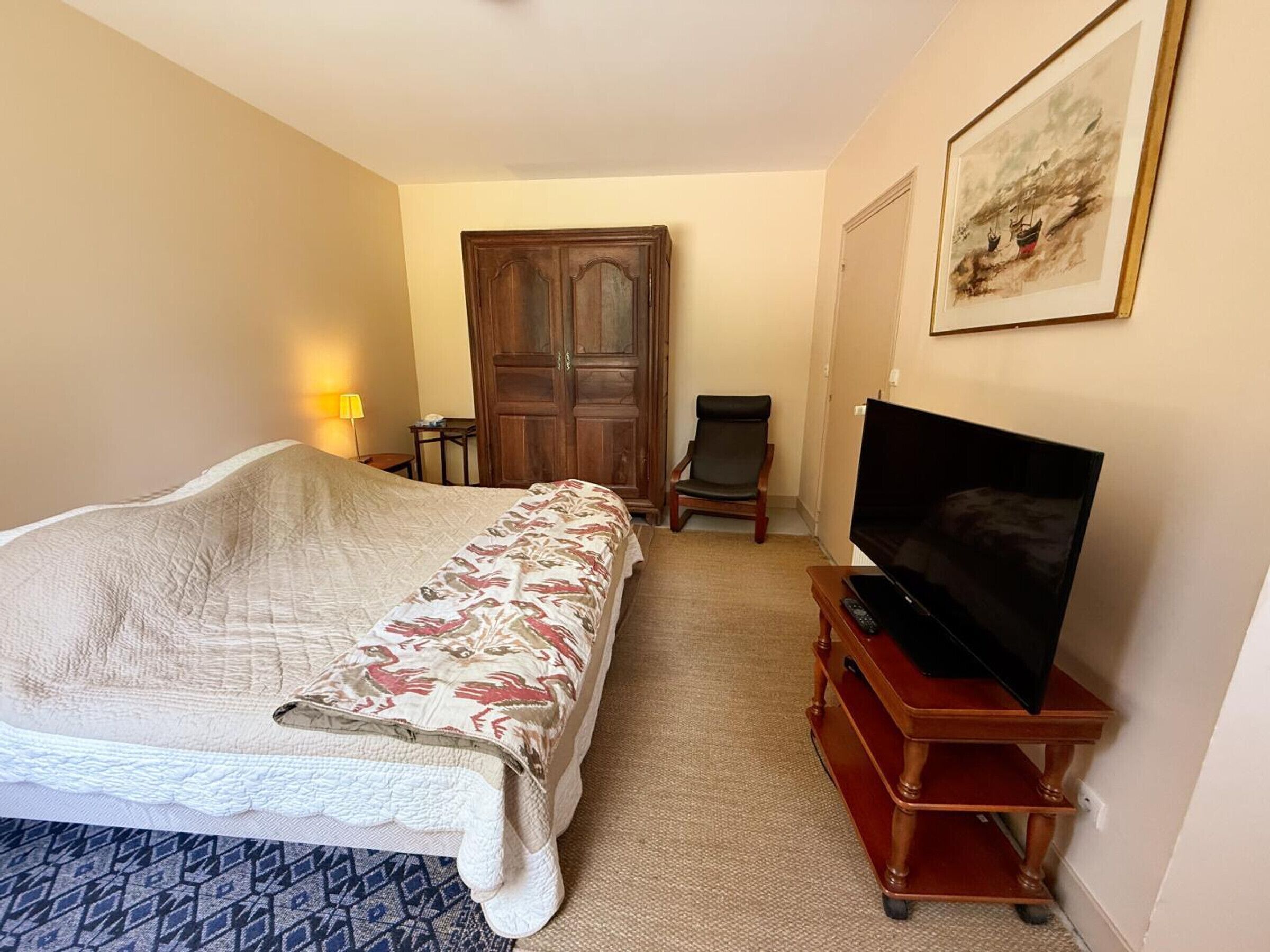 A bedroom with a bed, a chair, a TV on a wooden stand, a wardrobe, and a painting on the wall.
