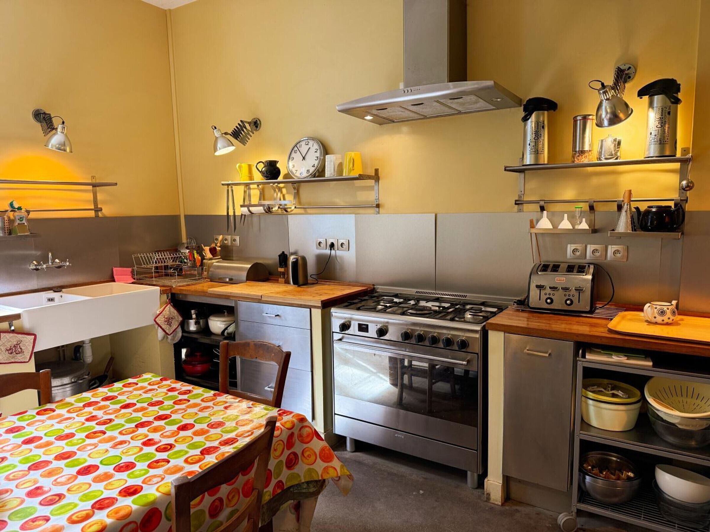 Une cuisine compacte équipée d’une cuisinière, d’un four et d’un grille-pain. On y trouve un évier, une table recouverte d’une nappe à pois et des étagères remplies d’ustensiles et d’appareils de cuisine.