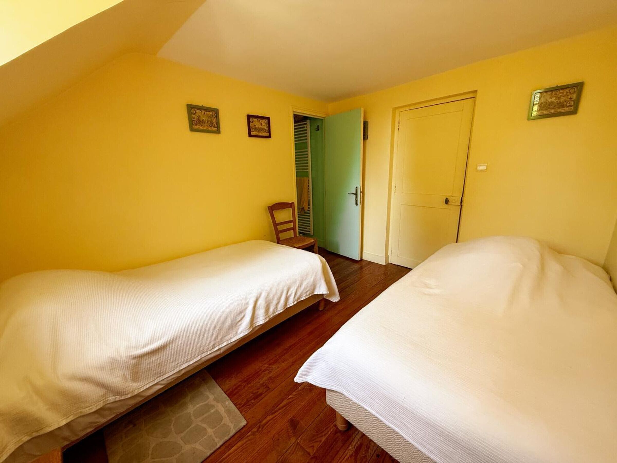 A room with two beds, wooden flooring, a door, and a chair.