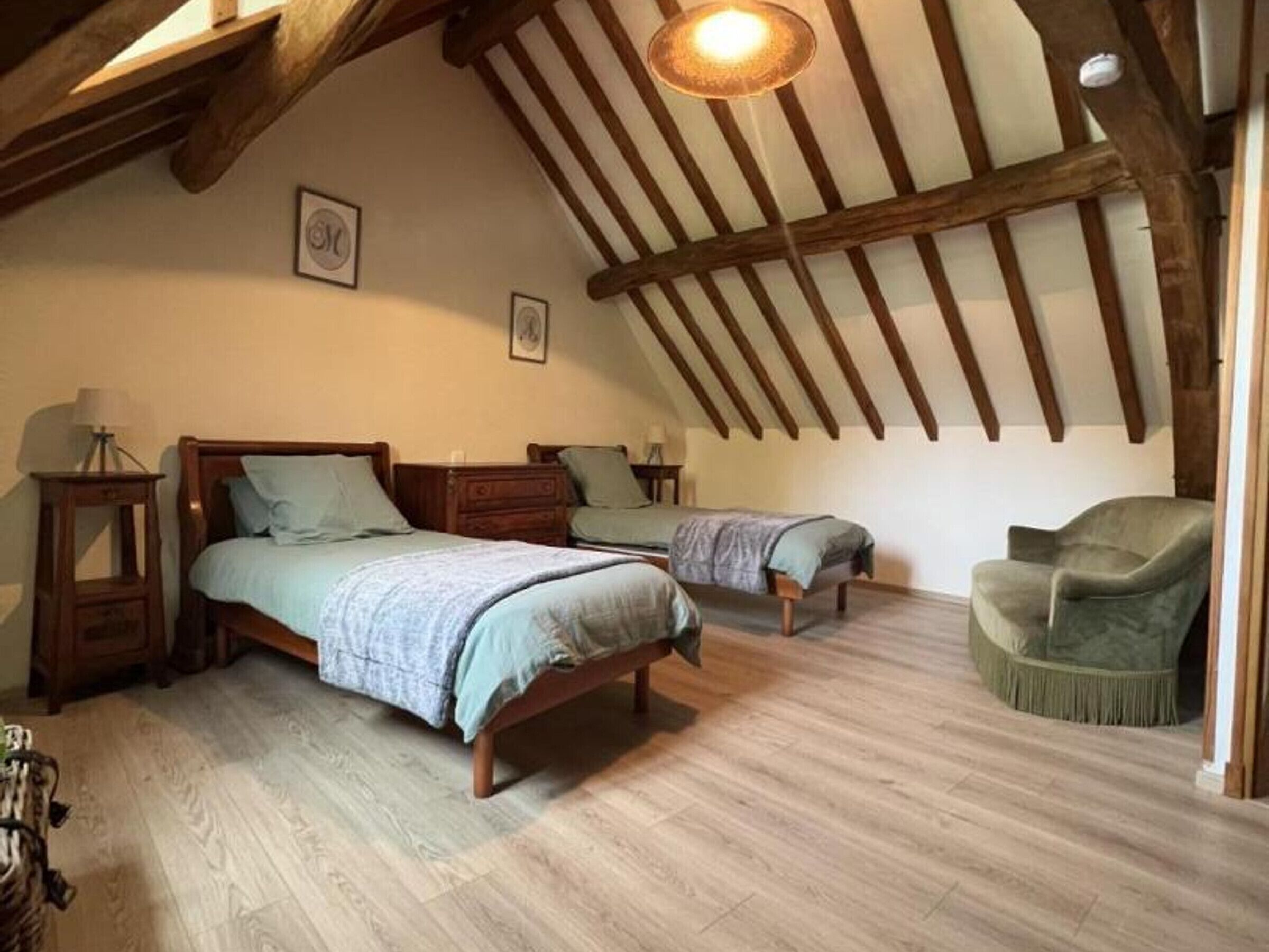 Une chambre avec deux lits, des poutres en bois, un luminaire suspendu et un fauteuil vert.