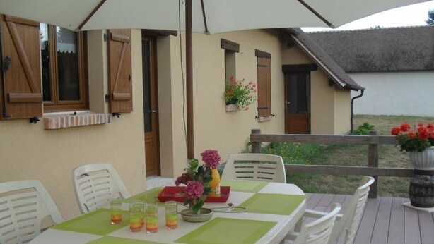 Un área de comedor al aire libre con una mesa preparada para cuatro personas, rodeada de sillas blancas. Hay una terraza de madera, una barandilla y vistas a un edificio con techo inclinado.