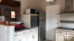 Cocina compacta con armarios blancos, microondas, horno y refrigerador. Hay un reloj en la pared y una lámpara colgante sobre la encimera.