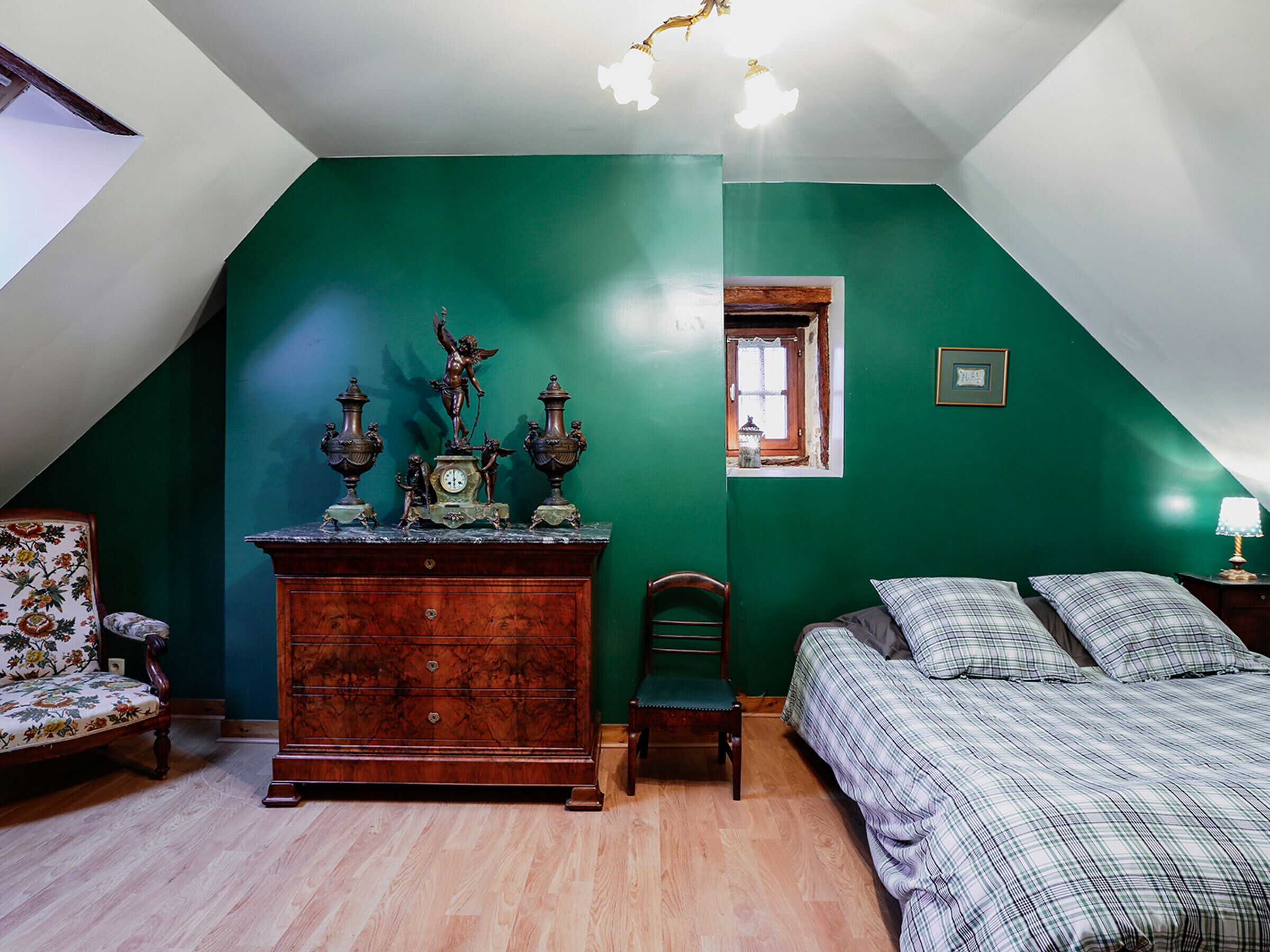 Ein Schlafzimmer mit schrägem Dach, einer Holzkommode mit Dekoration, einem Bett mit Karo-Bettwäsche und einem Stuhl.
