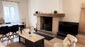 A living room with a fireplace, a TV, a dining area, and a kitchen visible in the background. - Cozy Renovated Cottage in Private Courtyard near Loire Castles (Avaray)