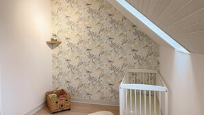 Una habitación con techo inclinado, una cuna, una pequeña cesta y una pared con diseño de hojas.