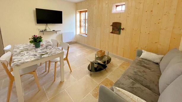 Una sala de estar con un sofá gris, una mesa de comedor con sillas, un televisor montado en la pared y una ventana con contraventanas de madera.