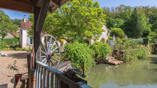 Un pont de bois rustique enjambant une rivière paisible, entouré d’une végétation luxuriante et de bâtiments traditionnels en pierre.